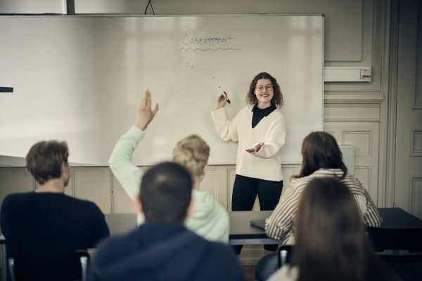 Lärare förklarar vid en whiteboard i klassrummet, en elev räcker upp handen.