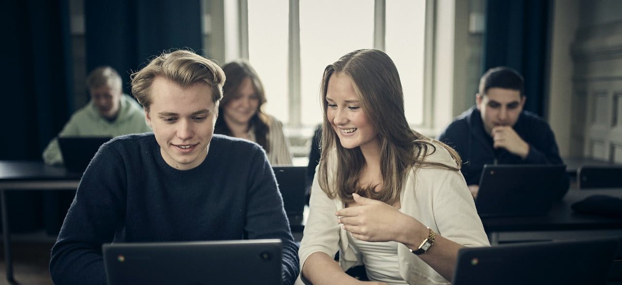 Två elever samarbetar och ler medan de tittar på en laptop i klassrummet.