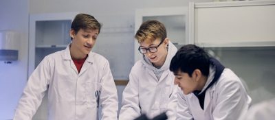 Tre elever med vita rockar under en lektion i naturkunskap