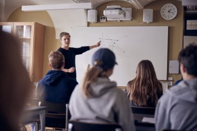 Lärare undervisar framme vid tavlan i ett klassrum.