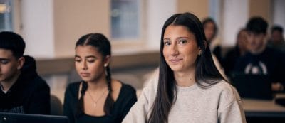 Elev sitter i ett klassrum med sin dator och kollar in i kameran.