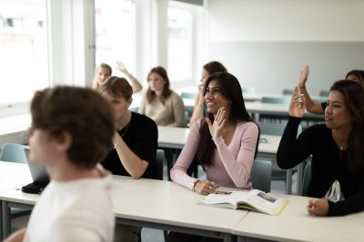 Elever sitter i ett klassrum och räcker upp sina händer