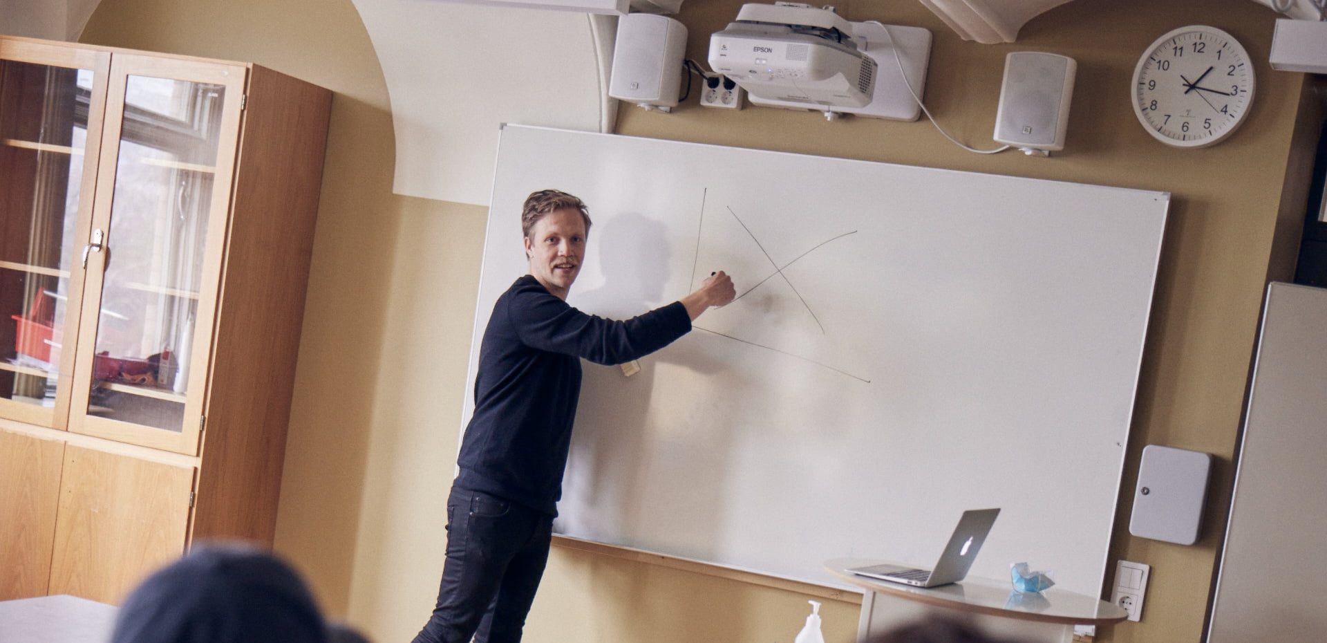 Lärare undervisar framme vid tavlan i ett klassrum.