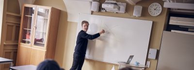 Lärare undervisar framme vid tavlan i ett klassrum.